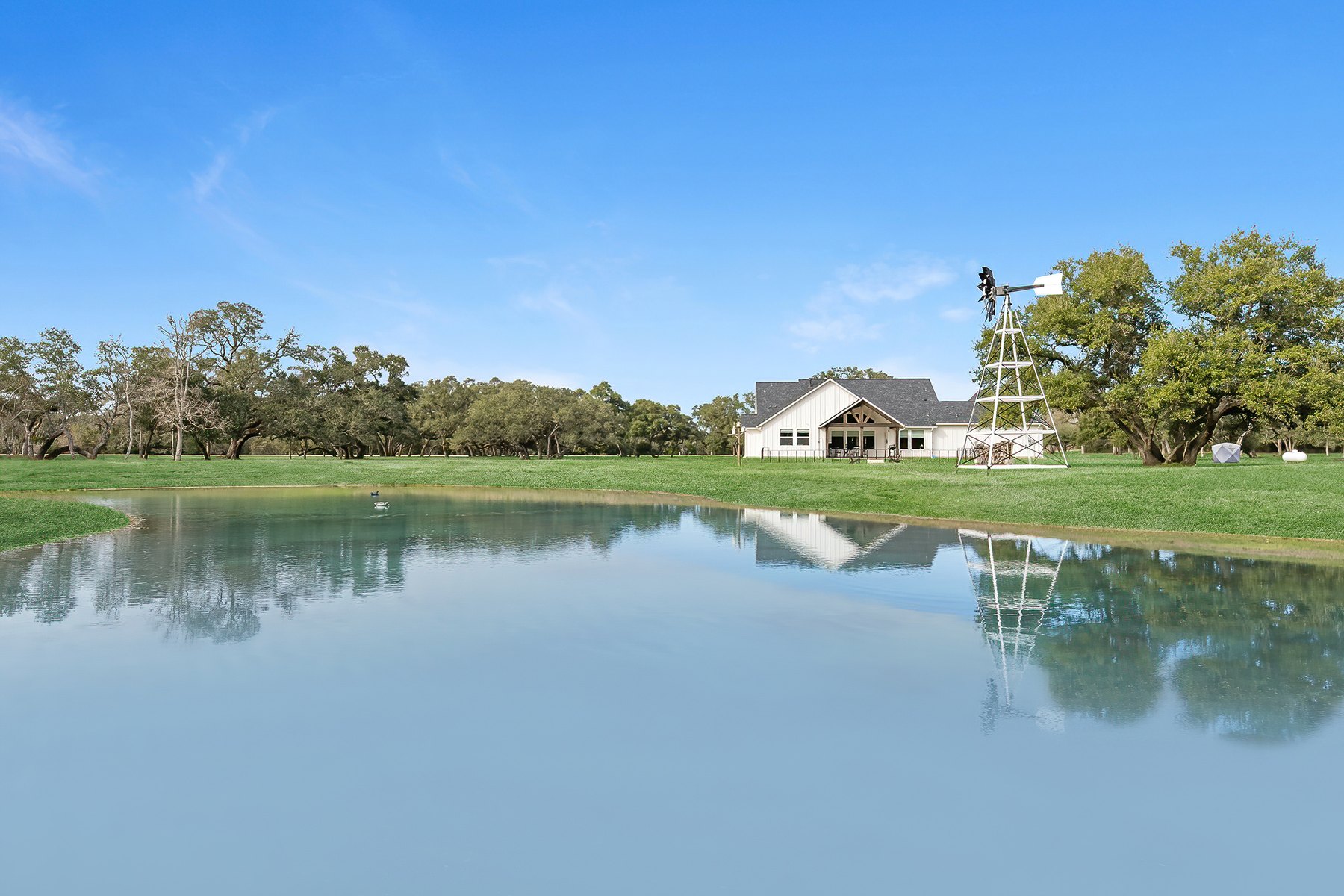 White farmhouse beside a pond with a windmill and live oaks